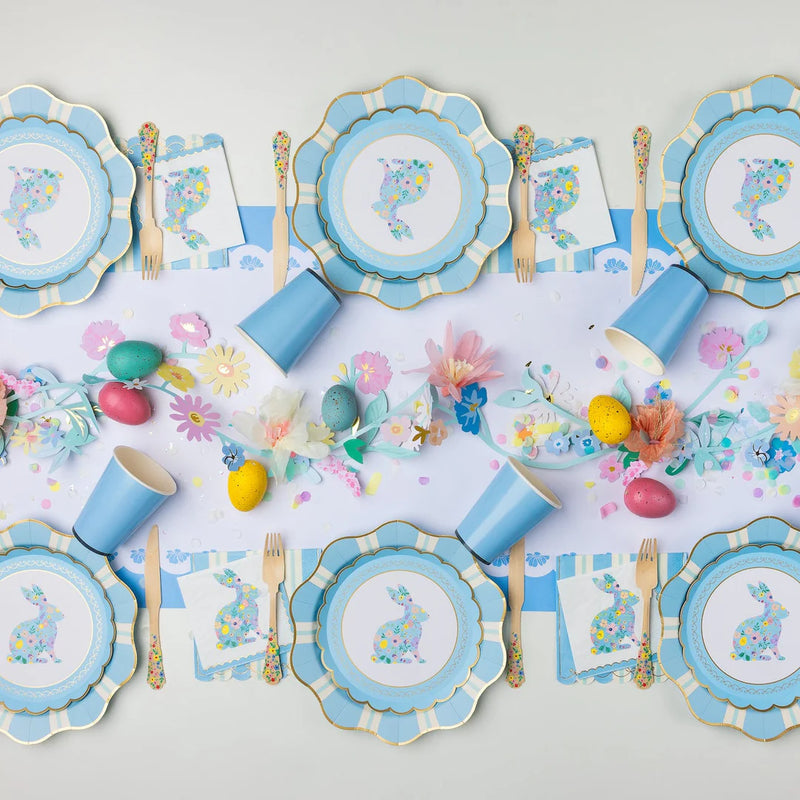 Blue striped scalloped paper guest napkins layered beneath floral bunny napkins on an Easter tablescape with blue scalloped paper plates, solid blue paper cups, floral wooden flatware, and a paper flower garland with speckled eggs.