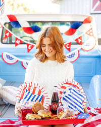 Lifestyle photo of woman holding a red tray loaded with burgers, blue striped star plates, red striped scalloped plates, and bottled sodas at an outdoor patriotic party
