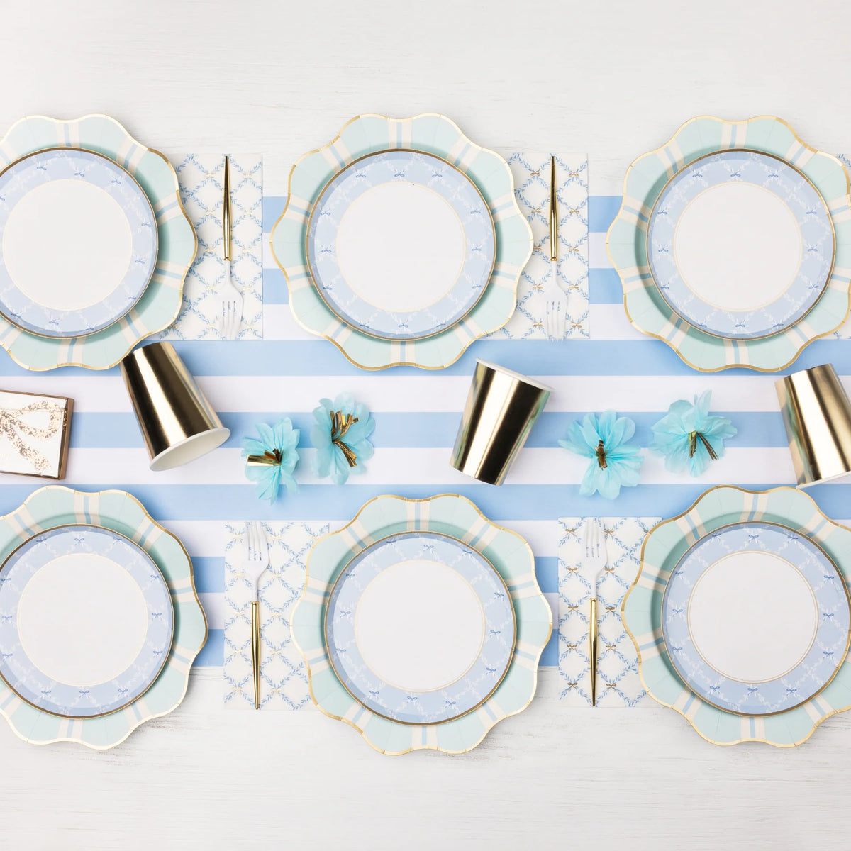 Blue trellis bow scalloped paper plates layered with mint and blue striped dinner plates on a blue and white striped runner with gold foil paper cups, gold flatware, and blue tissue paper flowers.