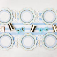 Blue trellis bow scalloped paper plates layered with mint and blue striped dinner plates on a blue and white striped runner with gold foil paper cups, gold flatware, and blue tissue paper flowers.