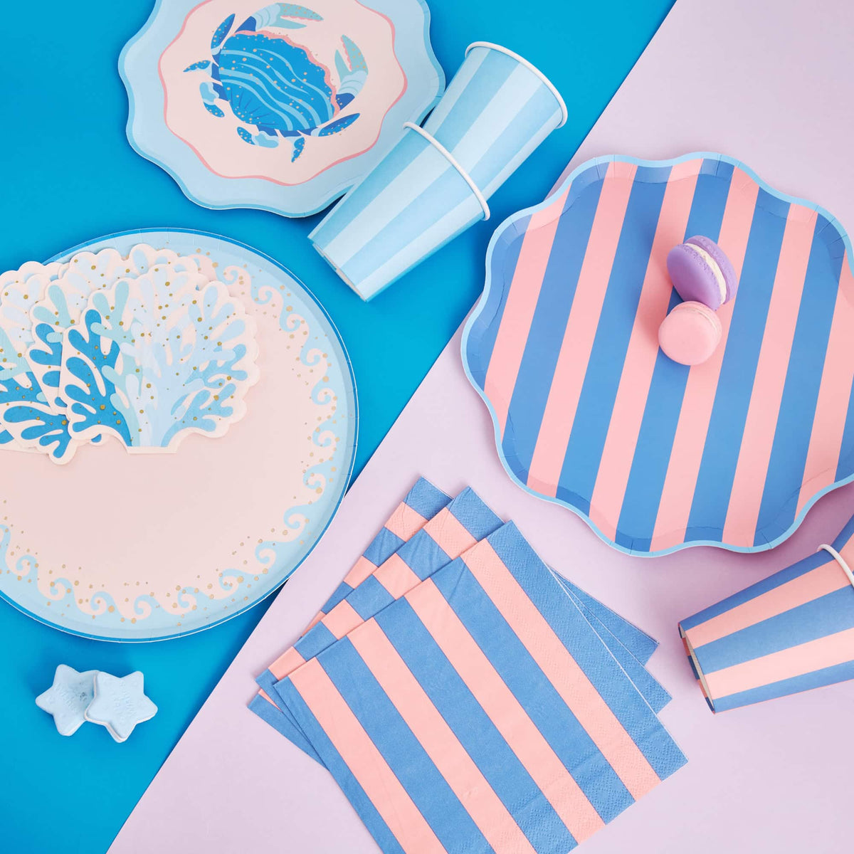 Coastal party flatlay with blue wave dinner plates, a crab plate, striped plates, coral napkins, cups, and macarons.