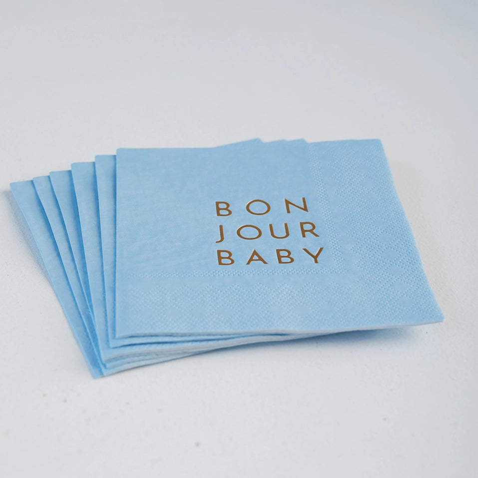 Stack of sky‑blue cocktail napkins with gold “BON JOUR BABY” lettering fanned on tabletop.