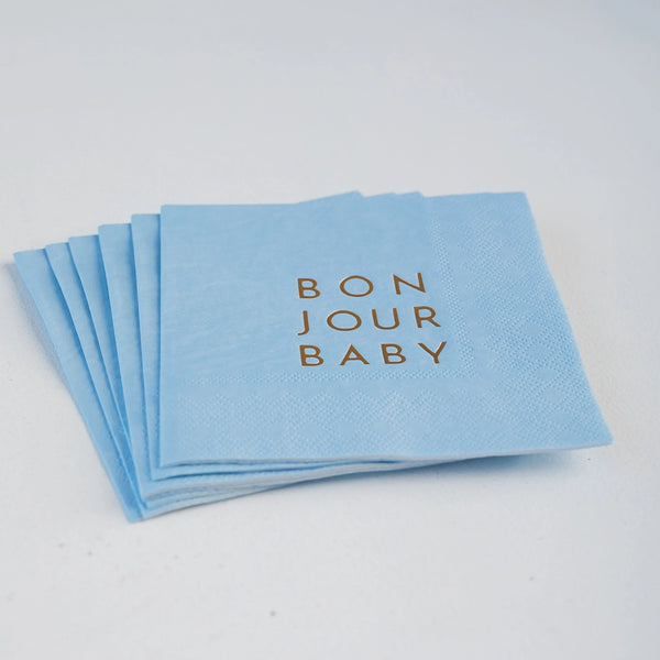 Stack of sky‑blue cocktail napkins with gold “BON JOUR BABY” lettering fanned on tabletop.