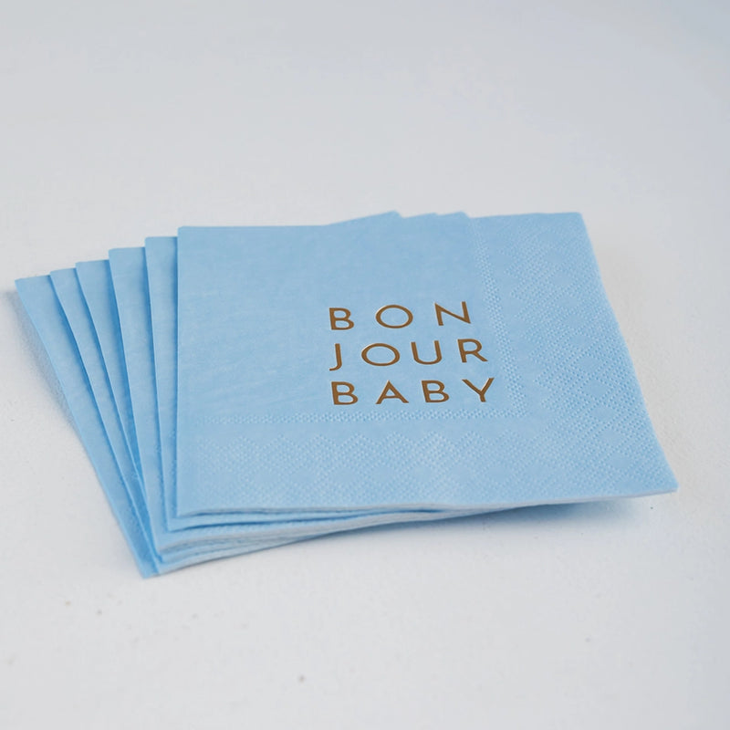 Stack of sky‑blue cocktail napkins with gold “BON JOUR BABY” lettering fanned on tabletop.