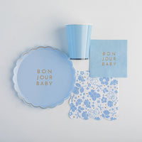 Flat‑lay of Bonjour Baby sky‑blue plate, cup, cocktail napkin and floral lunch napkin arranged on white background.