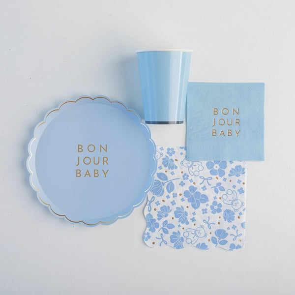 Flat‑lay of Bonjour Baby sky‑blue plate, cup, cocktail napkin and floral lunch napkin arranged on white background.