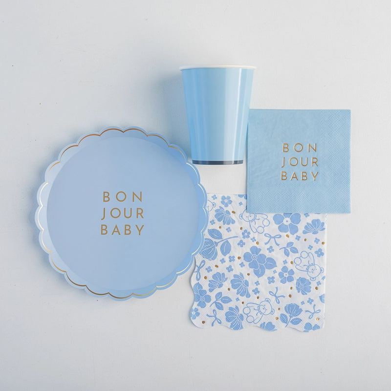 Flat‑lay of Bonjour Baby sky‑blue plate, cup, cocktail napkin and floral lunch napkin arranged on white background.