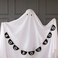 Whimsical scene of single tall sheet ghost posed against paneled wall; eyeholes cut reveal playful expression. Boo felt banner drapes in deep arc across the ghost’s front, starting high on left shoulder and cascading to right hip, repeating BOO five times with pom-pom spacers. Image illustrates banner curvature, lightweight construction and photo-booth potential, helping party planners visualize quick, camera-ready Halloween backdrop ideas.