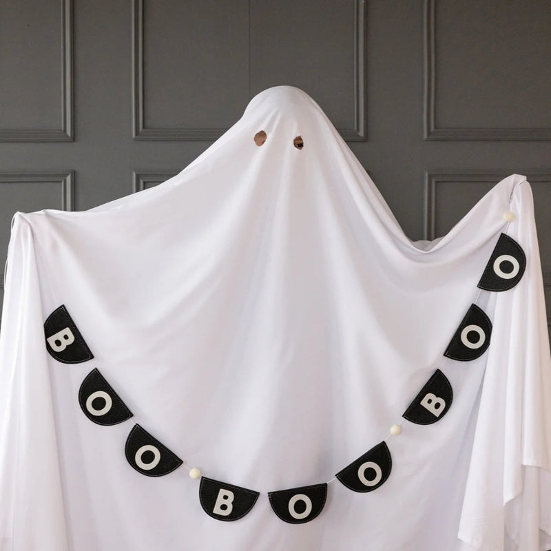Whimsical scene of single tall sheet ghost posed against paneled wall; eyeholes cut reveal playful expression. Boo felt banner drapes in deep arc across the ghost’s front, starting high on left shoulder and cascading to right hip, repeating BOO five times with pom-pom spacers. Image illustrates banner curvature, lightweight construction and photo-booth potential, helping party planners visualize quick, camera-ready Halloween backdrop ideas.