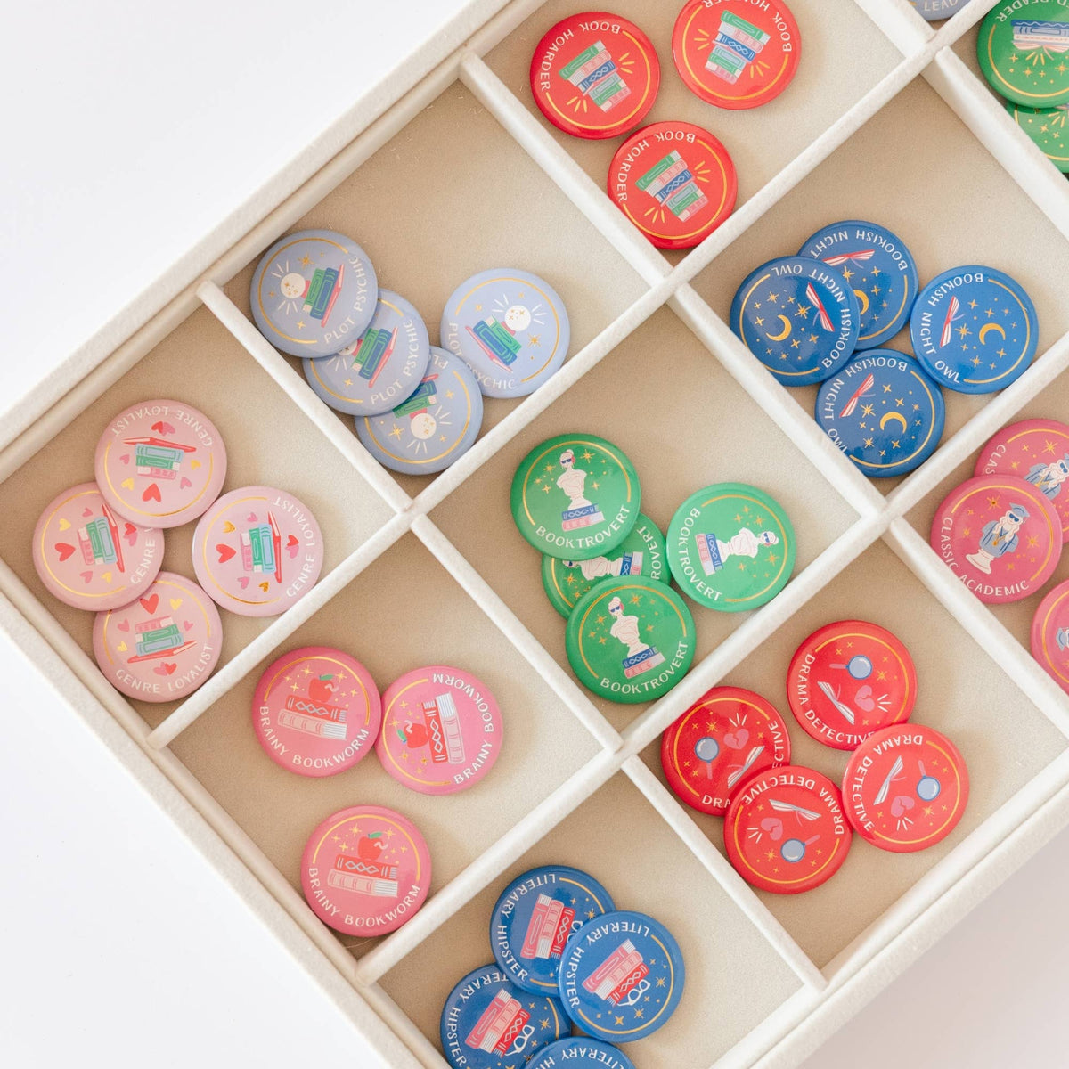 Jollity Co. Book Club Merit Badge pin buttons arranged in compartmentalized organizer trays sorted by design — pink Genre Loyalist and Brainy Bookworm, blue Bookish Night Owl, lavender Plot Psychic, green Booktrovert, red Drama Detective and Book Hoarder, and dark blue Literary Hipster pin buttons