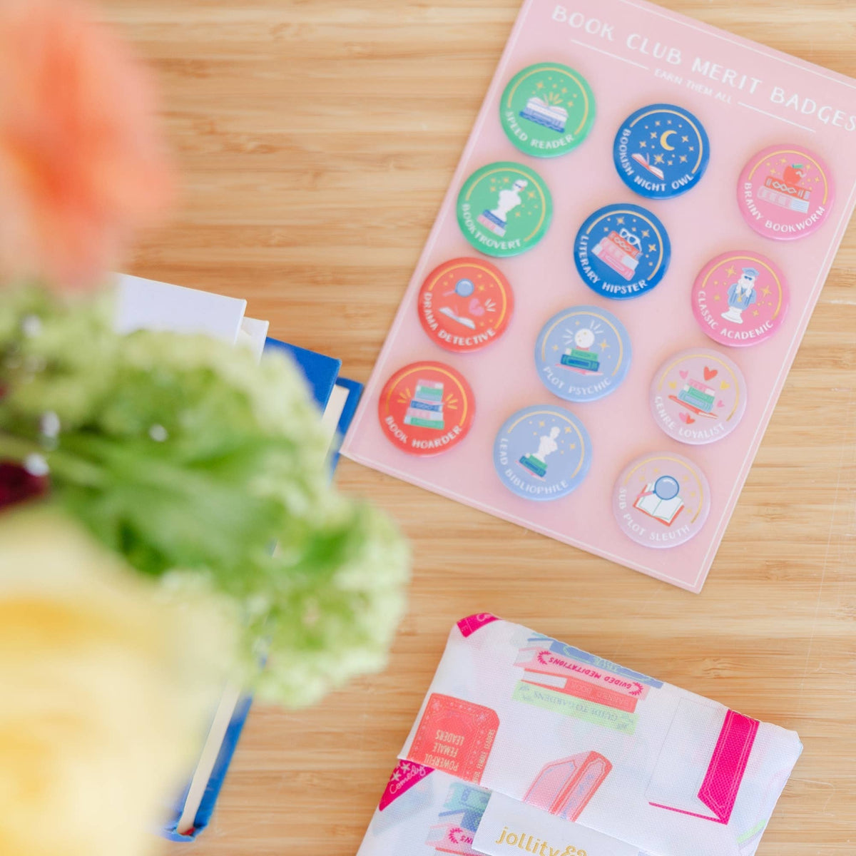 Book Club Merit Badges card with 12 pin-back buttons displayed on a wooden table alongside a Jollity Co. printed tote bag, a navy book, and out-of-focus flowers and hydrangeas