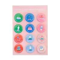 Book Club Merit Badges display card showing 12 unique pin-back button designs in a 3-by-4 grid on a soft pink background, with "Book Club Merit Badges — Earn Them All" lettering at the top