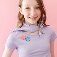 Smiling tween girl with braces wearing a lavender ribbed shirt with a lavender scarf, displaying a red Drama Detective merit badge and a blue Bookish Night Owl merit badge pinned to her shirt, photographed against a pink background