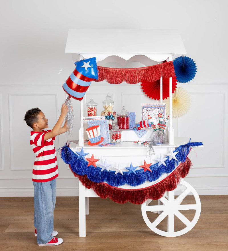 Boy in a red and white striped shirt holding a patriotic firecracker shaped foil balloon with silver fringe tail next to a decorated 4th of July party cart