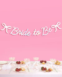 Bride to Be iridescent bow banner hung above bridal shower table with champagne coupes and desserts