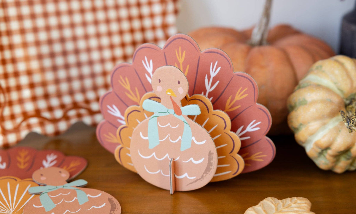 3D paper turkey decoration on a wooden surface with pumpkins and a checkered cloth in the background.