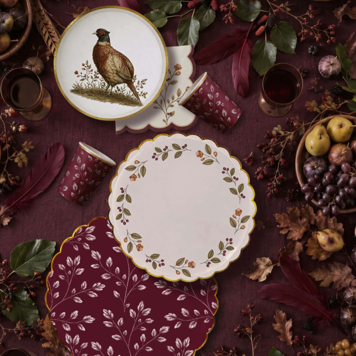 Two burgundy leaf cups on plum linen among feathers, pears, grapes, and dark fall foliage
