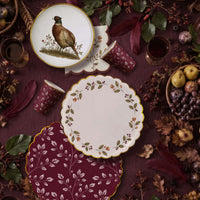 Two burgundy leaf cups on plum linen among feathers, pears, grapes, and dark fall foliage