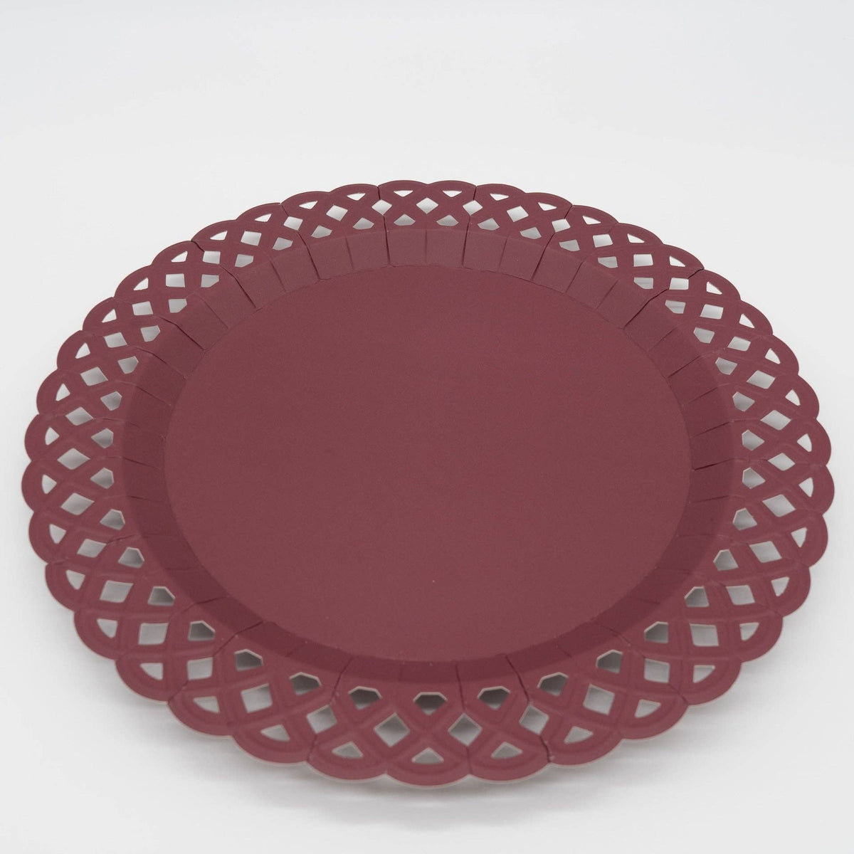 Angled close-up of a deep burgundy paper dinner plate showing the solid wine-colored center, raised rim, and intricate woven lattice cutout border.
