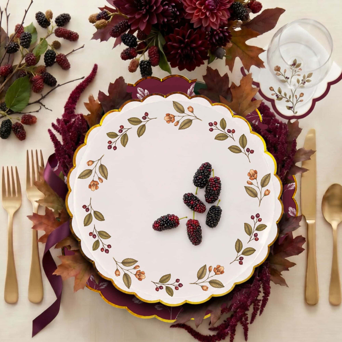 Overhead burgundy place setting with a berry wreath plate, gold flatware, dark dahlias, and a berry napkin under a wine glass