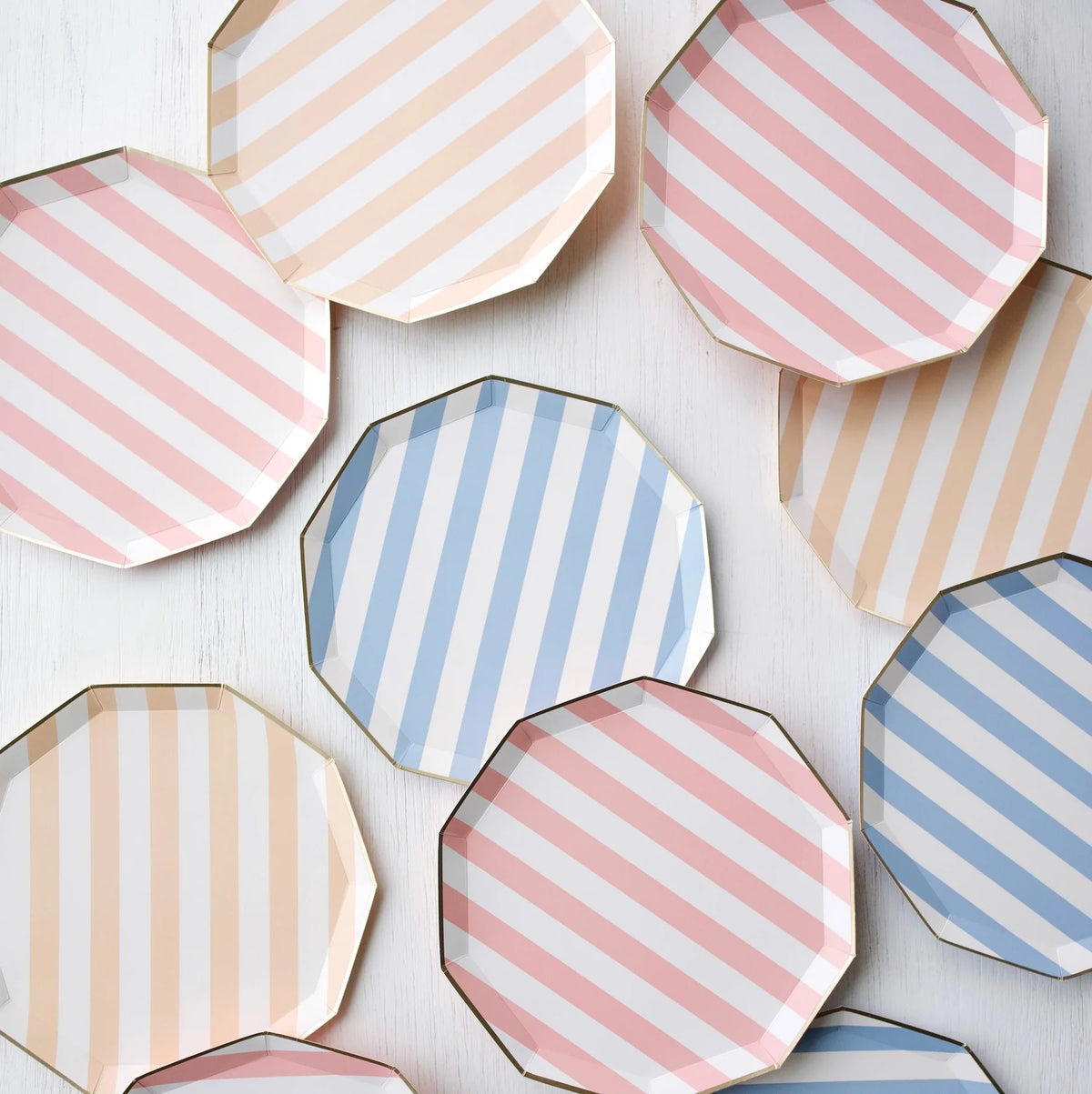 Cabana stripe paper plates in pink, peach, and sky blue colorways scattered across a white wood surface