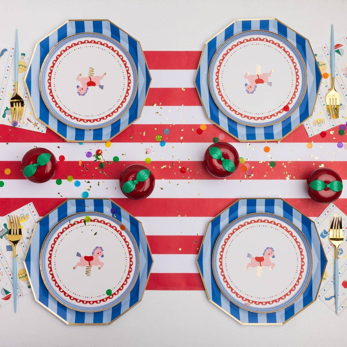 Overhead first birthday tablescape with blue and white striped octagonal paper dinner plates layered with Petite Playground carousel horse paper cake plates, Petite Playground lunch napkins, gold cutlery with light blue handles, red and white striped table runner, and red apple-shaped place card holders on a confetti-scattered surface