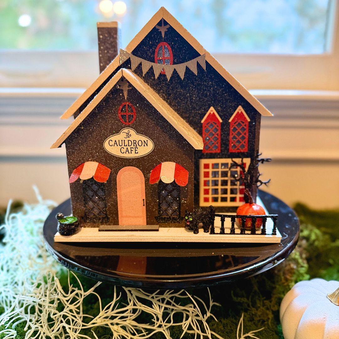 Vintage Halloween Putz house featuring The Cauldron Café with glowing windows, red accents, black cat, pumpkin, and spooky glitter details.