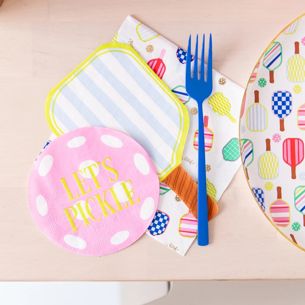 Blue fork resting on layered paddle guest napkins (lime‑stripe, pink pickleball, multicolor paddles) next to matching dinner plate on pale wood table.