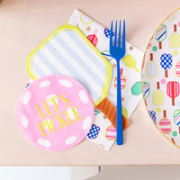 Blue fork resting on layered paddle guest napkins (lime‑stripe, pink pickleball, multicolor paddles) next to matching dinner plate on pale wood table.