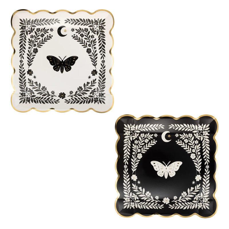 Two square paper plates with gold scalloped edges, one cream with a black moth and black floral border, one black with a cream moth and cream floral border, each with a crescent moon and stars