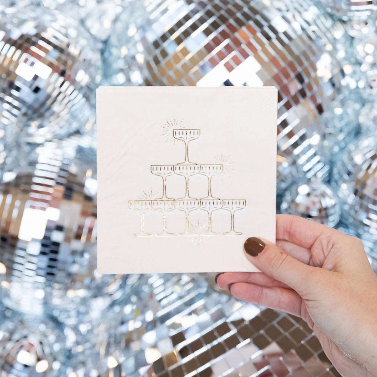 Hand holding a blush champagne tower cocktail napkin with gold foil coupe pyramid design against a silver disco ball backdrop for a New Year's Eve party