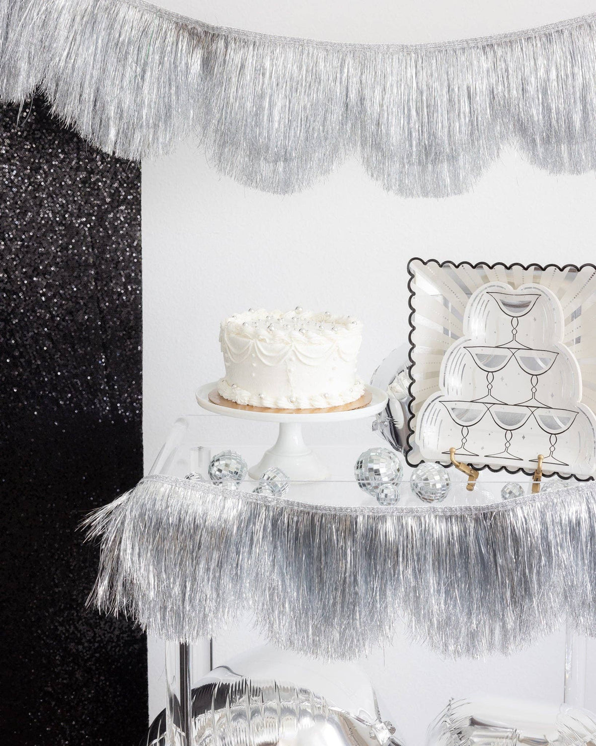 Champagne tower shaped plates styled on a party table with a cake and silver fringe backdrop