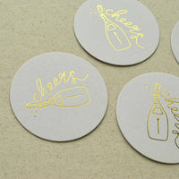 Round white Champagne Coaster with gold foil champagne bottle and cheers script.