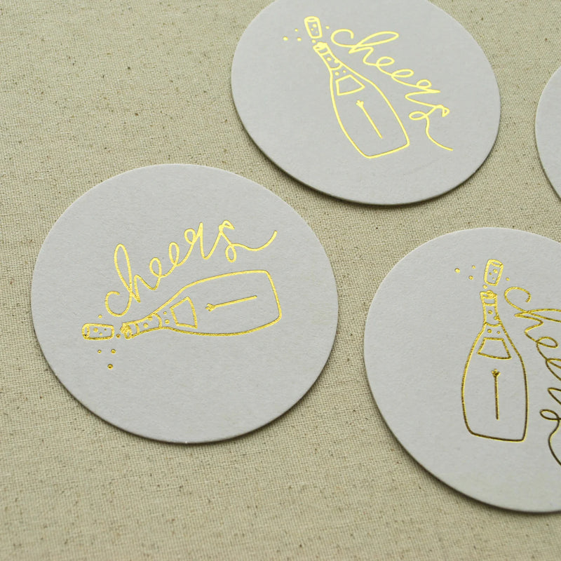 Round white Champagne Coaster with gold foil champagne bottle and cheers script.