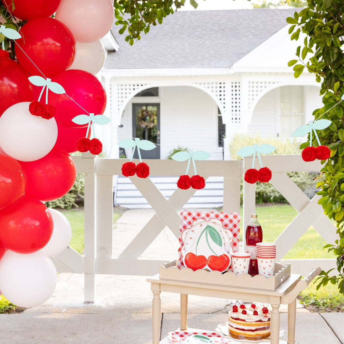 Cherry pom pom banner displayed near red and white balloons at a cherry themed party setup.
