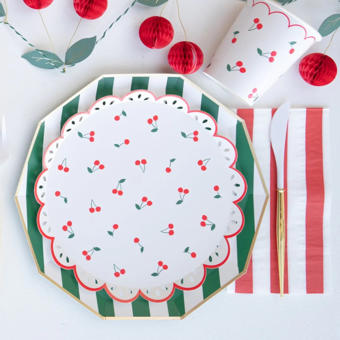 Cherry print paper plate layered on a green striped dinner plate with a red and white striped guest towel napkin, gold flatware, and cherry print paper cups.