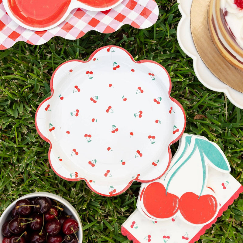 Cherry themed picnic setup with cherry plates, napkins, and dessert on grass.
