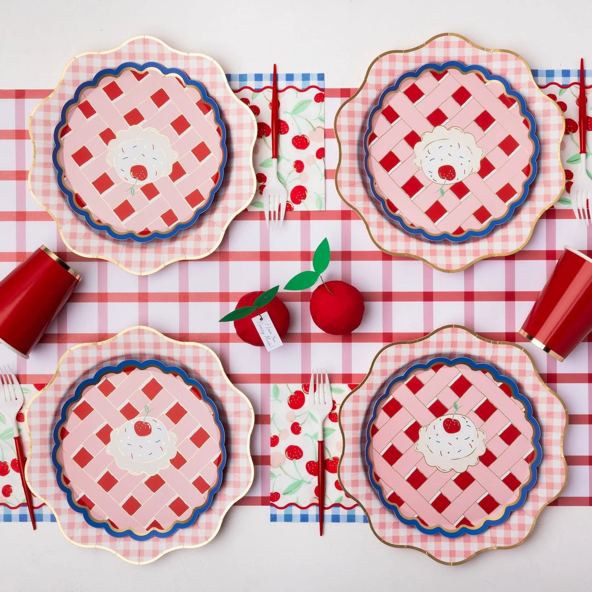 Overhead picnic table for four with cherry print lunch napkins, cherry pie paper plates, red cups, and gingham table runners.
