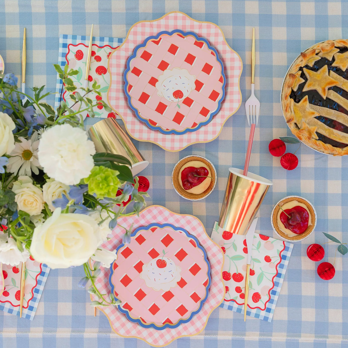 Overhead summer cherry pie tablescape on a blue gingham cloth with cherry print lunch napkins, pie-shaped plates, gold cups, and a star-topped lattice pie.