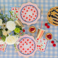 Overhead summer cherry pie tablescape on a blue gingham cloth with cherry print lunch napkins, pie-shaped plates, gold cups, and a star-topped lattice pie.