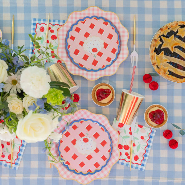 Overhead summer cherry pie tablescape on a blue gingham cloth with cherry print lunch napkins, pie-shaped plates, gold cups, and a star-topped lattice pie.