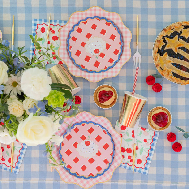 Overhead summer cherry pie tablescape on a blue gingham cloth with cherry print lunch napkins, pie-shaped plates, gold cups, and a star-topped lattice pie.