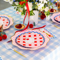 Cherry print paper lunch napkin styled at a place setting with a pie-shaped plate, gold cutlery, and blue gingham linens.