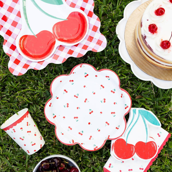 Cherry themed party picnic setup with cherry plates, shaped napkins, cake, and cherries on grass.
