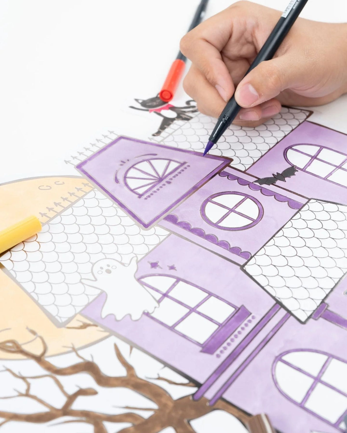 Vertical crop showing a child's hand coloring the haunted house paper placemat with purple and orange markers.