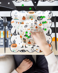 Close-up of a child’s hand picking green candy from the patterned bowl atop a matching plate on a striped table runner.