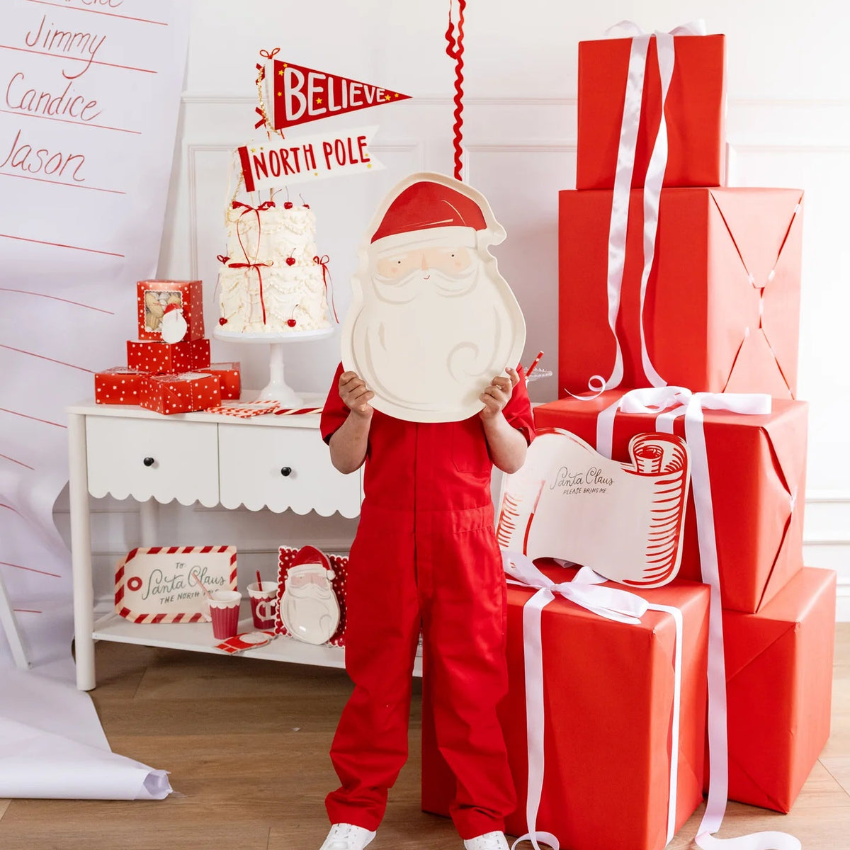Full shot of a child standing behind the Santa tray next to a large stack of Christmas presents.