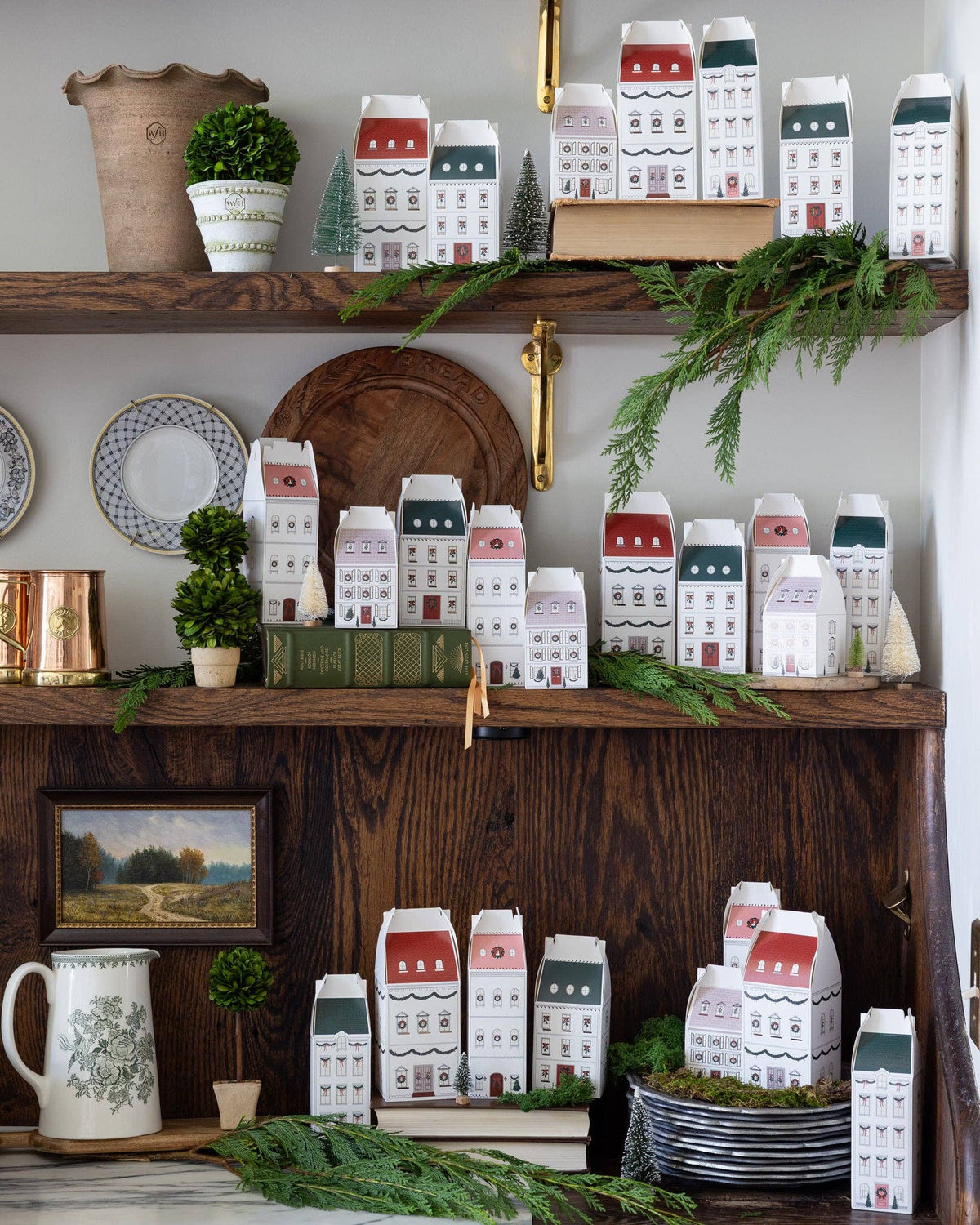 Rows of house-shaped Christmas advent boxes on wooden kitchen shelves with bottle brush trees and cedar garland