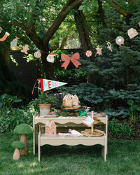 Christmas icon paper garland strung between trees above an outdoor treat table; MERRY felt pennant, gingerbread house boxes, and napkins shown separately, not included