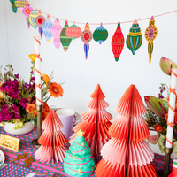 Styled holiday scene showing the ornament garland hung above pastel fabric Christmas trees, candy canes, and colorful holiday décor.